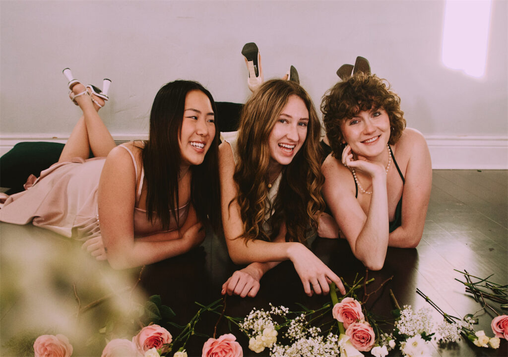 Three girls are wearing AW Bridal bridesmaid dresses and laughing happily in the room. There are also some flowers on the ground.