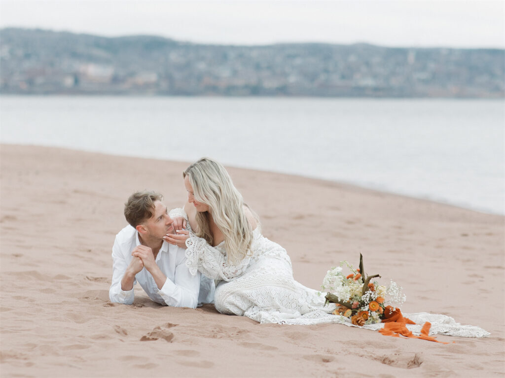 A couple lying on the beach, with the woman gently touching the man's face.