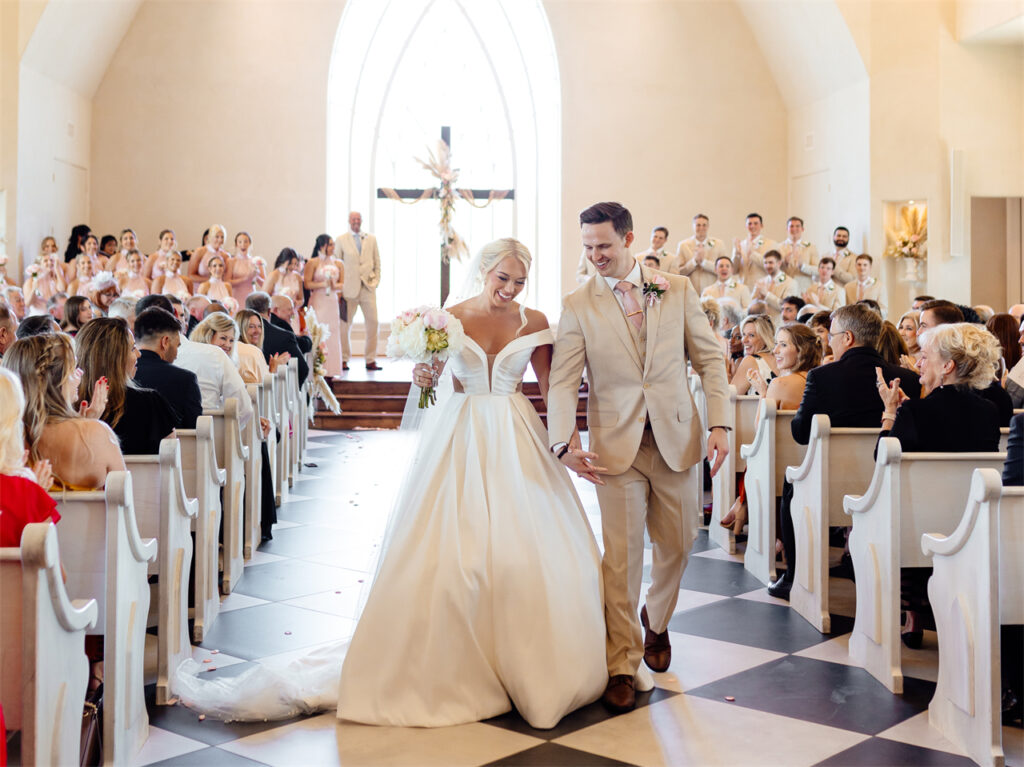 A newlywed couple walks down the aisle in a church, smiling and holding hands, surrounded by applauding guests.